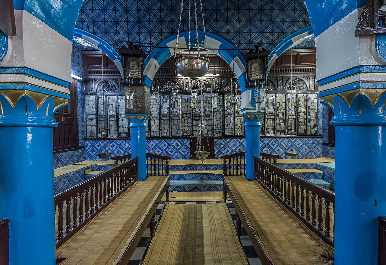Wnętrze synagogi z niebieskimi kafelkami, kolumnami i drewnianymi ławami.