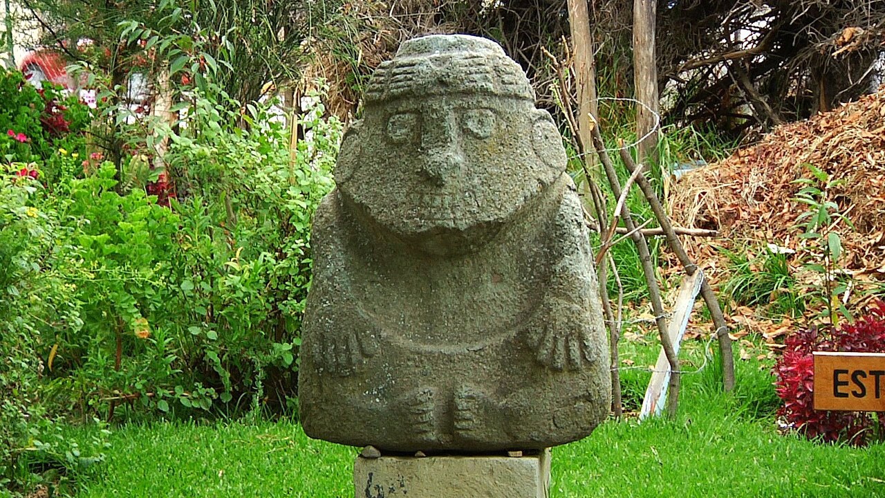 Kamienna figura kultury Recuay w ogrodzie Muzeum Archeologicznego Áncash: postać z dłońmi złożonymi na piersi, w opasce na głowie.